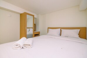 A hotel room with a bed, a wooden headboard, a nightstand, a mirror, and two rolled towels. - Homey And Comfort Studio Loft Apple 1 Condovilla Apartment (Jakarta)