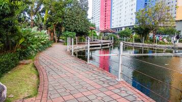 A paved walkway beside a water body with a bridge, surrounded by greenery and tall buildings in the background.