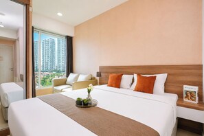 A hotel room with a large bed, a sofa, a small table with a vase, and a view of high-rise buildings. - Chic Minimalist Studio At Apartment Grand Kamala Lagoon (Bekasi)