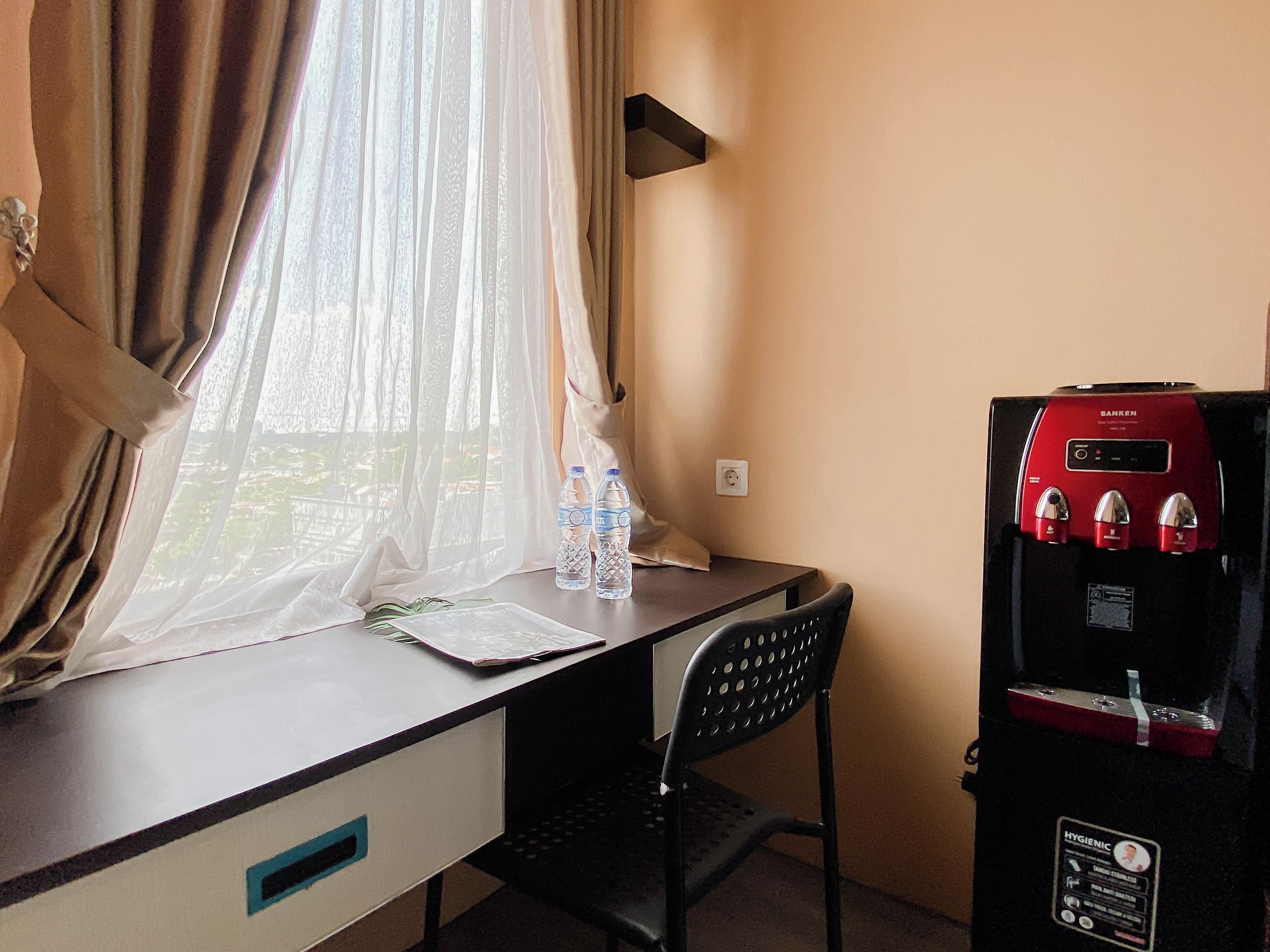 A hotel room with a desk, a chair, a window with sheer curtains, and a minibar with a water dispenser.