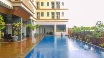 A swimming pool with a tiled deck, surrounded by potted plants and a multi-story building with balconies.