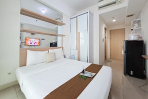 A hotel room with a bed, a television, a refrigerator, and a door leading to another room. - Neat And Nice Studio Room At Apartment Tree Park City Bsd (Tangerang)