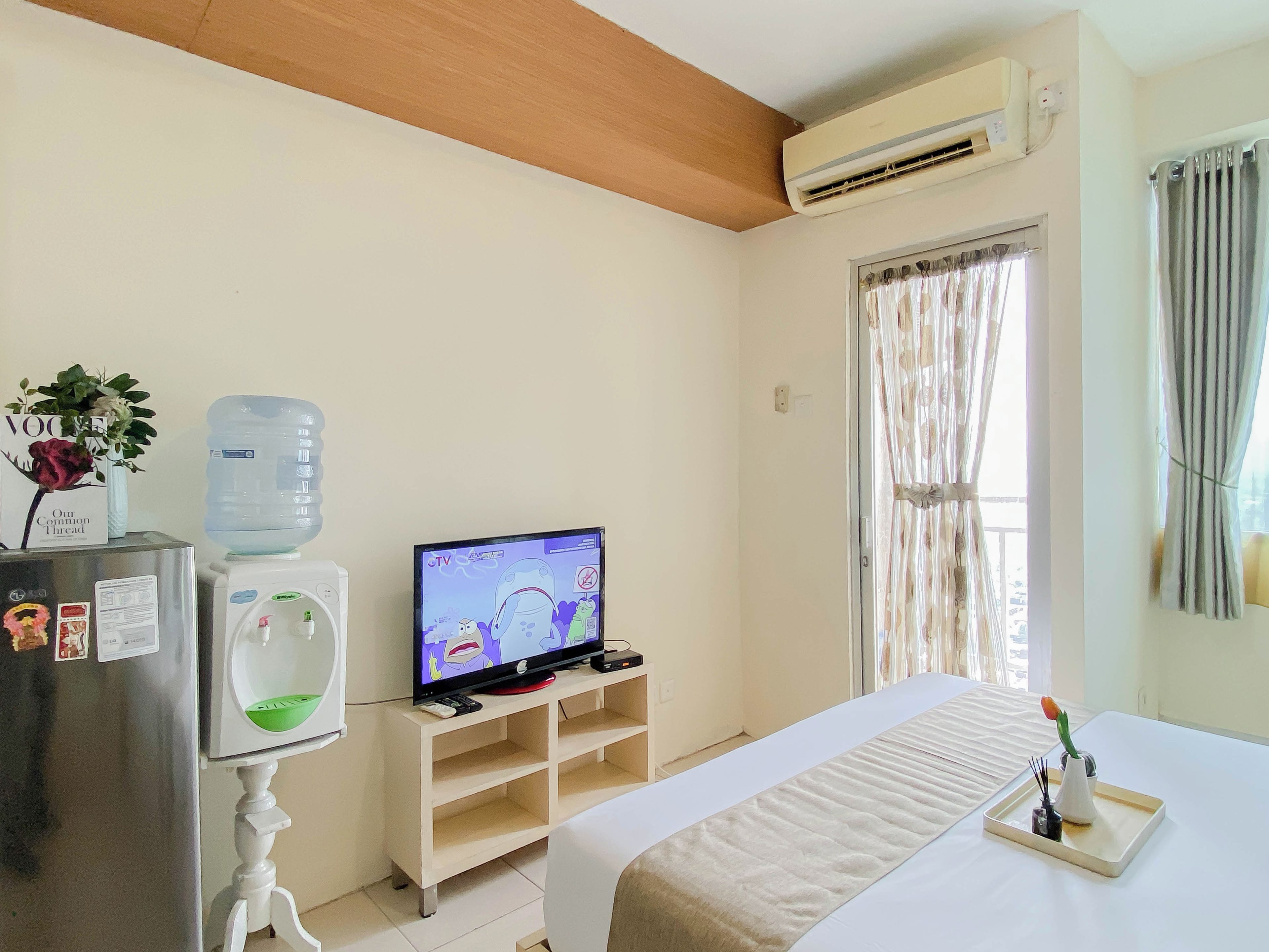 A hotel room with a bed, a TV, a water dispenser, a refrigerator, and a window with curtains.