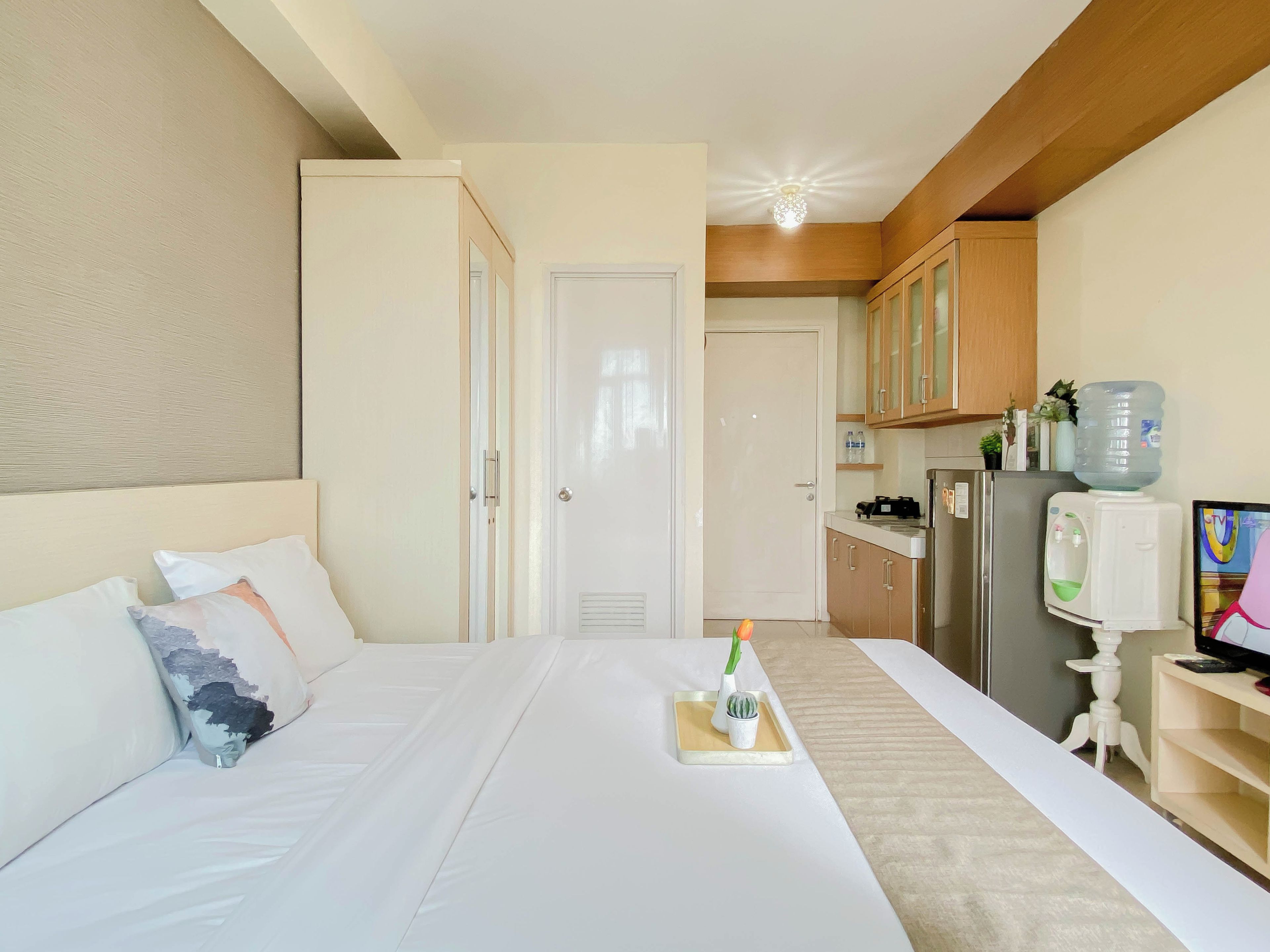 A hotel room with a bed, bedside table, a small plant, a water dispenser, and a refrigerator.