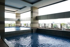 A modern indoor swimming pool with a city view, surrounded by a minimalist design and large windows. - Elegant 2Br Loft Apartment Maqna Residence (Jakarta)