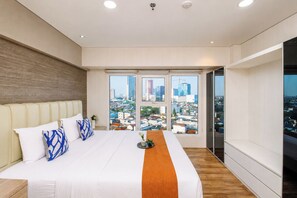 A modern bedroom with a large bed, a wooden headboard, and a city view through floor-to-ceiling windows. - Elegant 2Br Loft Apartment Maqna Residence (Jakarta)
