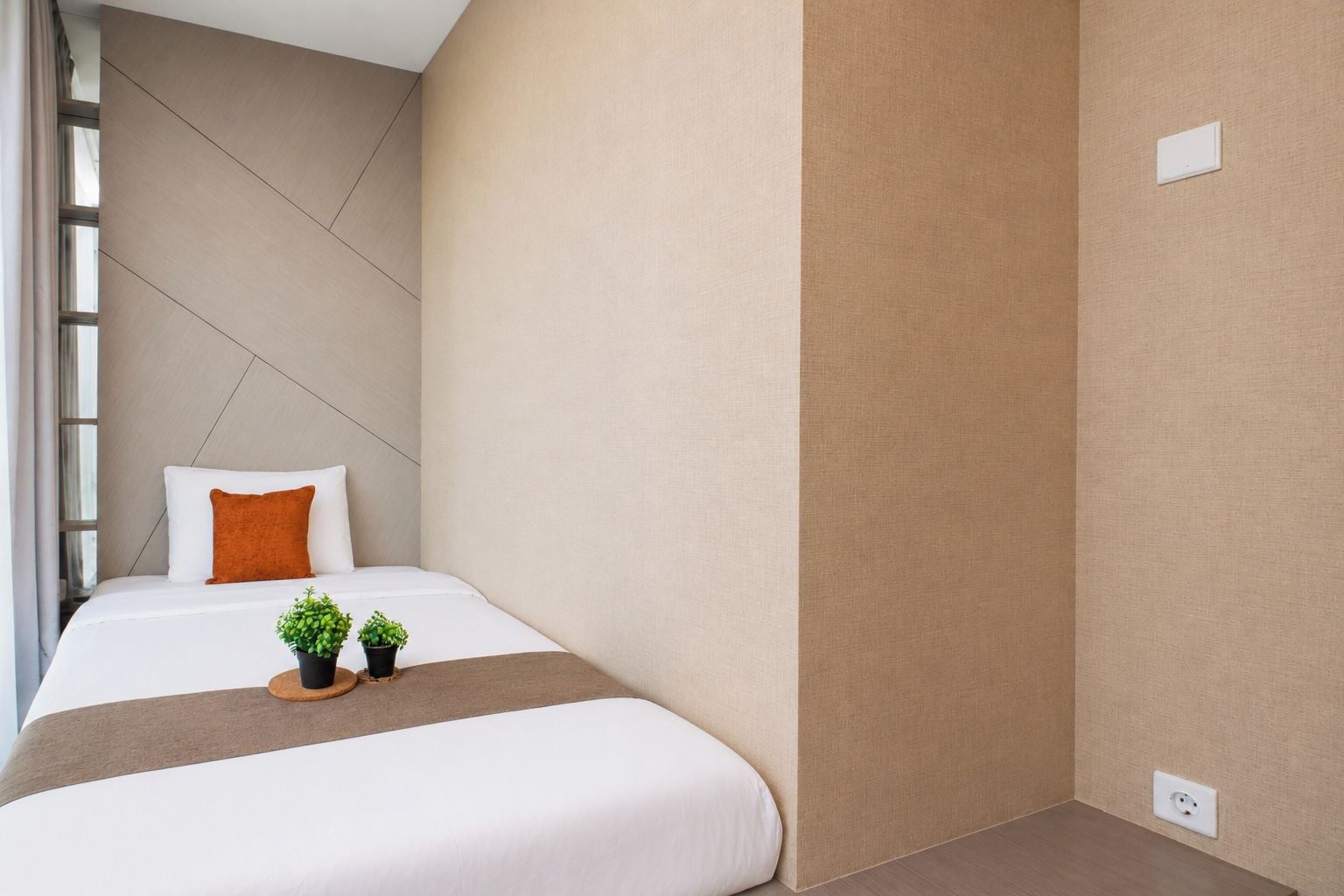 Habitación de hotel con una cama, dos plantas en macetas, una ventana y un interruptor de luz en la pared.