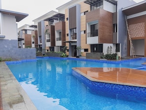 A modern residential building with a swimming pool, featuring a combination of brick and white facades, and a wooden deck area. - Homey And Roomy 3Br At Apartment Asatti (Tangerang)