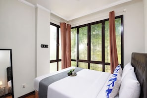 A hotel room with a bed, a window with curtains, a bedside table with a lamp, and a wall-mounted control panel. - Homey And Roomy 3Br At Apartment Asatti (Tangerang)