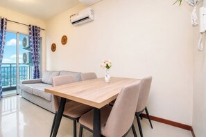 A modern living room with a wooden dining table, beige sofa, and a view of the city through large windows. - Simple And Cozy 1Br At Apartment Green Bay Condominium (Jakarta)