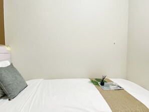 Ein Einzelbett mit weißer Matratze, einem grauen Kissen, einem beigen Nachttisch und einem kleinen schwarzen Gegenstand auf dem Tisch.