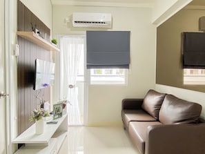 A living room with a brown leather sofa, a mounted TV, a white air conditioning unit, and a window with a view of buildings outside. - Good Homey 2Br At Apartment Green Pramuka City (Jakarta)