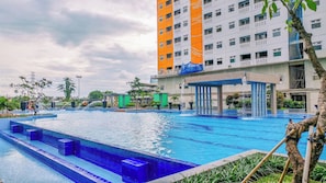 A swimming pool with a water fountain, surrounded by greenery and a multi-story building with balconies. - Good Homey 2Br At Apartment Green Pramuka City (Jakarta)