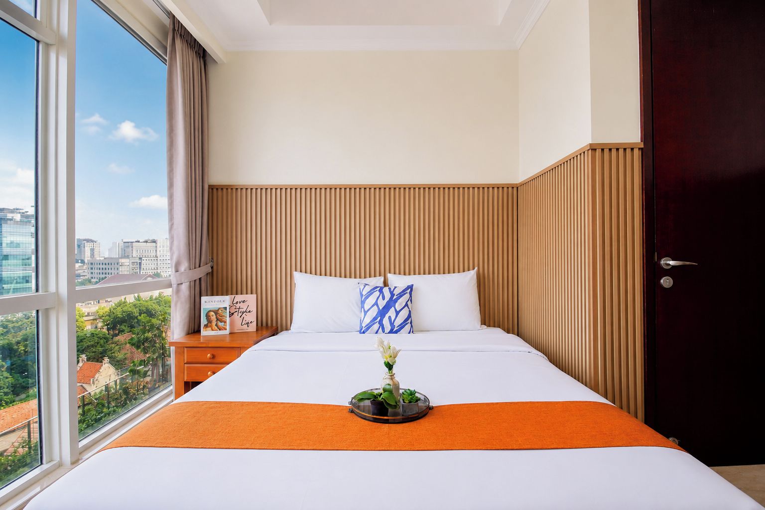 Habitación de hotel con un ventanal grande, una cama con ropa blanca, un plaid naranja, una mesita de noche con un libro y una puerta de madera oscura.