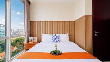 Habitación de hotel con un ventanal grande, una cama con ropa blanca, un plaid naranja, una mesita de noche con un libro y una puerta de madera oscura.