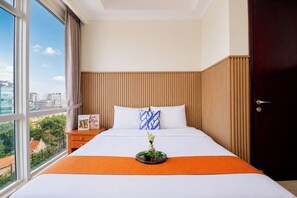 Habitación de hotel con un ventanal grande, una cama con ropa blanca, un plaid naranja, una mesita de noche con un libro y una puerta de madera oscura.