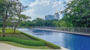 A swimming pool with a wooden deck, surrounded by greenery and a metal fence. - Serene 1Br With Sky View At Apartment Izzara (Jakarta)
