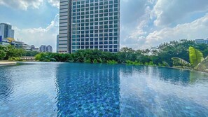 A swimming pool with a tiled edge, surrounded by greenery and a tall building in the background. - Serene 1Br With Sky View At Apartment Izzara (Jakarta)