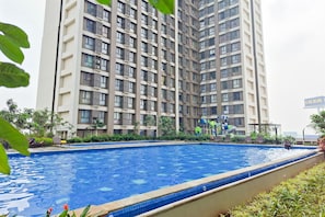 A swimming pool with a tiled edge, surrounded by greenery and a high-rise building in the background. - Neat And Cozy Studio At Apartment Sky House Alam Sutera (Tangerang)
