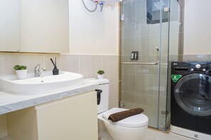 A modern bathroom with a white sink, two small potted plants, a glass shower enclosure, and a black washing machine. - Serene 3Br With Working Room At Apartment Meikarta District 1 (Cikarang)