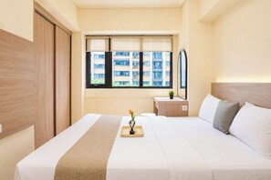 A hotel room with a large bed, a bedside table, a window with blinds, and a view of a building outside. - Serene 3Br With Working Room At Apartment Meikarta District 1 (Cikarang)