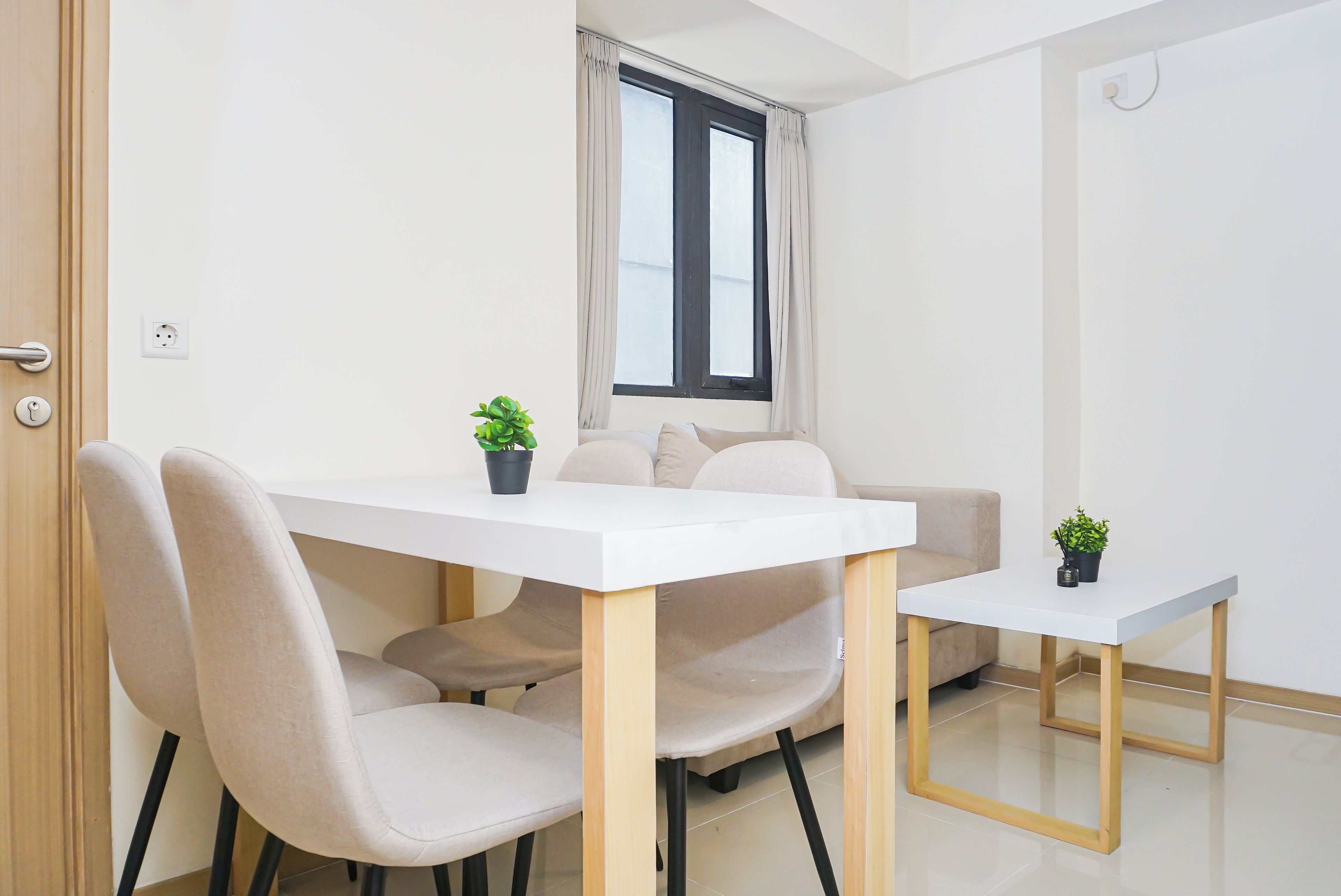 Una habitación pequeña y moderna con una mesa blanca y dos sillas, un sofá, una ventana con cortinas y una planta en maceta sobre la mesa.