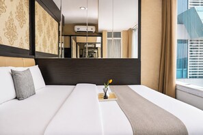 A hotel room with a bed, a bedside table with a vase of flowers, and a window with curtains. - Functional And Cozy Studio At Apartment Grand Kamala Lagoon (Bekasi)