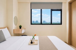 Ein Hotelzimmer mit einem Bett, einem Nachttisch mit Pflanze, einem Fenster mit Blick auf die Stadt und einer Tür.