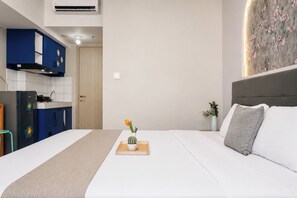 A hotel room with a bed, a bedside table with a vase, a kitchenette with blue cabinets, and a door. - Functional Homey Studio Room At Apartment Springlake Summarecon Bekasi (Bekasi)
