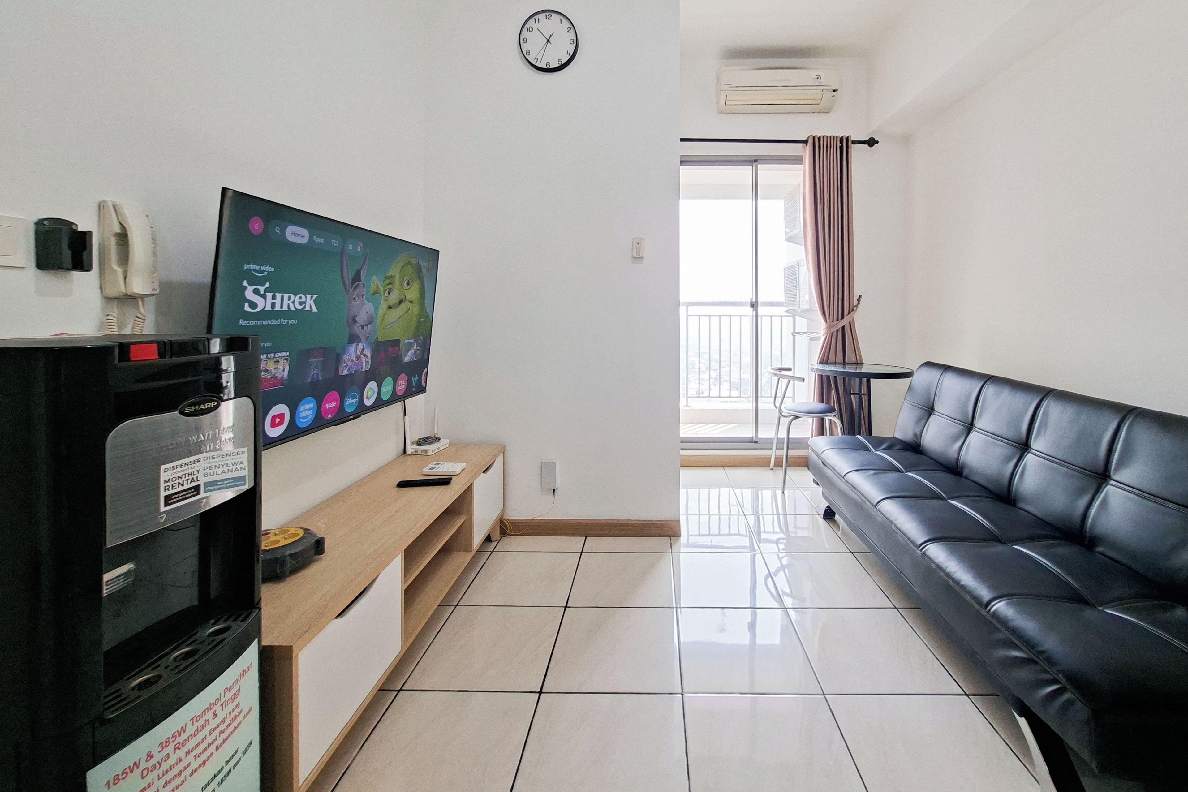 Sala de estar com sofá de couro preto, televisão de tela plana na parede, mesa de centro de madeira e uma pequena mesa com uma cadeira ao fundo.