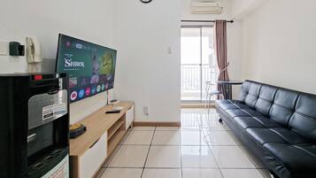 Sala de estar com sofá de couro preto, televisão de tela plana na parede, mesa de centro de madeira e uma pequena mesa com uma cadeira ao fundo.