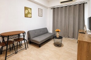 Ein Wohnzimmer mit grauem Sofa, einem runden Holztisch mit Stühlen, einem kleinen schwarzen Beistelltisch mit Blumenvase und einem Fernseher auf einem Holzgestell.