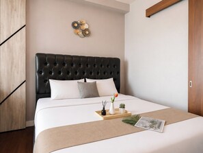 A neatly made bed with a tufted headboard, a bedside table with a tray holding a plant and a book, and a wall-mounted decorative piece. - Homey Modern 1Br At Apartment Grand Kamala Lagoon (Bekasi)