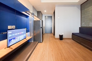 Une chambre d’hôtel moderne avec un téléviseur à écran plat posé sur une étagère en bois, un réfrigérateur intégré, un plancher en bois et un canapé de couleur foncée.