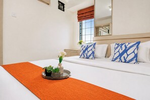 A hotel room with a bed, a bedside table, a mirror, and a window with blinds. - Cozy Strategic 3Br At Apartment Bassura City Near Shopping Mall (Jakarta)