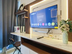 A modern living room with a flat-screen TV displaying a streaming service interface, a remote control, and a vase with flowers on the counter. - Restful Studio Room At Tamansari Iswara Apartment (Bekasi)