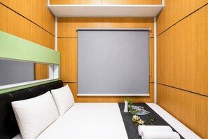 Une chambre d’hôtel avec un lit, une fenêtre avec un store, une table de chevet avec un vase et un cadre photo, et un mur vert.