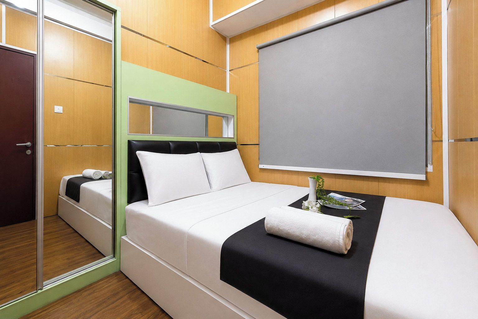 A hotel room with a bed, a bedside table, a mirror, a window with a blind, and a door.