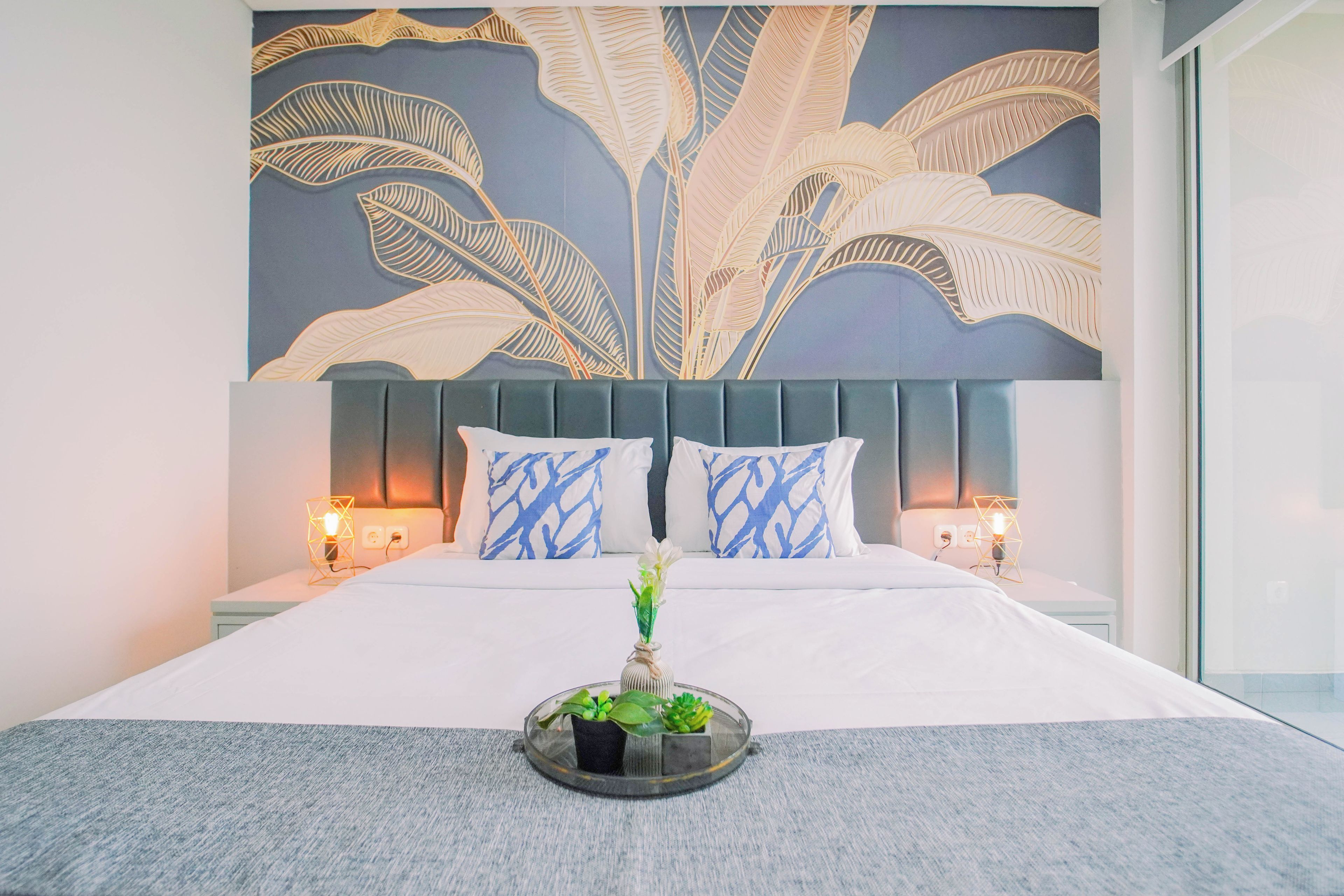 A hotel room with a large bed, two bedside lamps, a decorative wall mural, and a bedside plant.