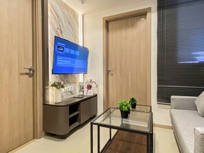 A modern living room with a flat-screen TV mounted on the wall, a glass coffee table, a sofa, and a wooden cabinet with decorative items. - Versatile Smart 2Br At Apartment Meikarta District 2 (Cikarang)