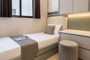 A compact bedroom with a single bed, a bedside table, a small stool, and a window with curtains. - Versatile Smart 2Br At Apartment Meikarta District 2 (Cikarang)