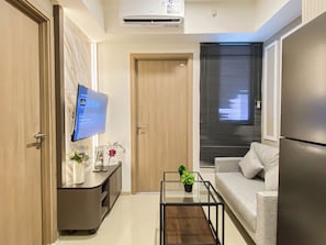A modern living room with a flat-screen TV, a sofa, a coffee table, and a refrigerator. - Versatile Smart 2Br At Apartment Meikarta District 2 (Cikarang)