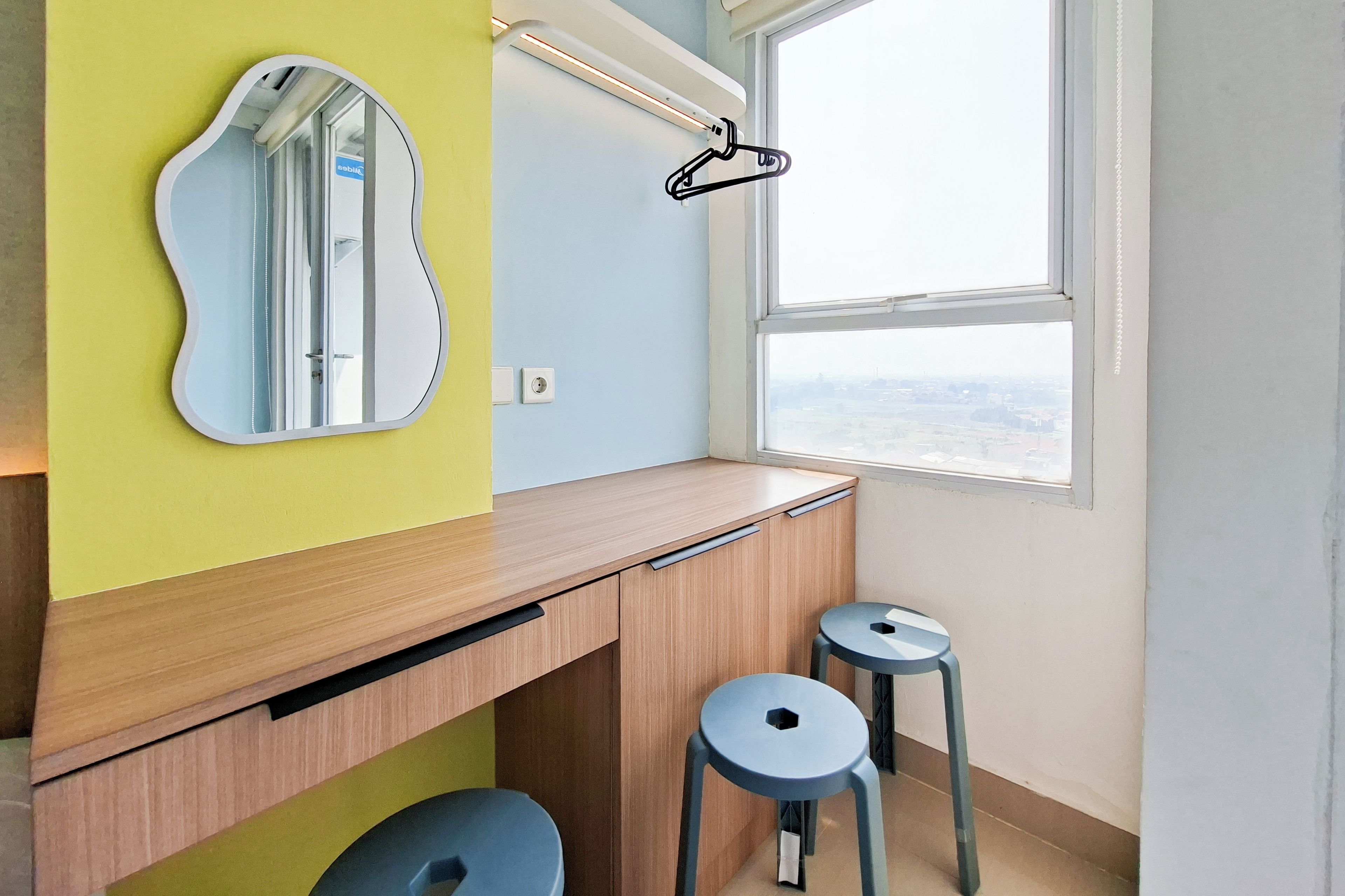 A small, brightly colored room with a wooden desk, two stools, a mirror, and a window with a view.