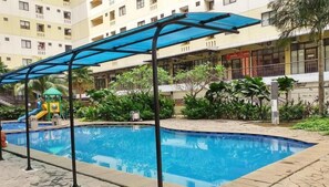 A swimming pool with a blue canopy, surrounded by greenery and a playground. - Tranquil And Modern Studio At Apartment Kebagusan City (Jakarta)