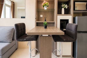 A modern kitchen with a wooden table, two bar stools, a sofa, and a refrigerator. - Homey And Warm 1Br At Apartment Vasanta Innopark (Cikarang)