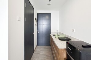 Um corredor estreito com uma porta escura, uma pequena cozinha com pia e bancada, e uma mala preta à direita.