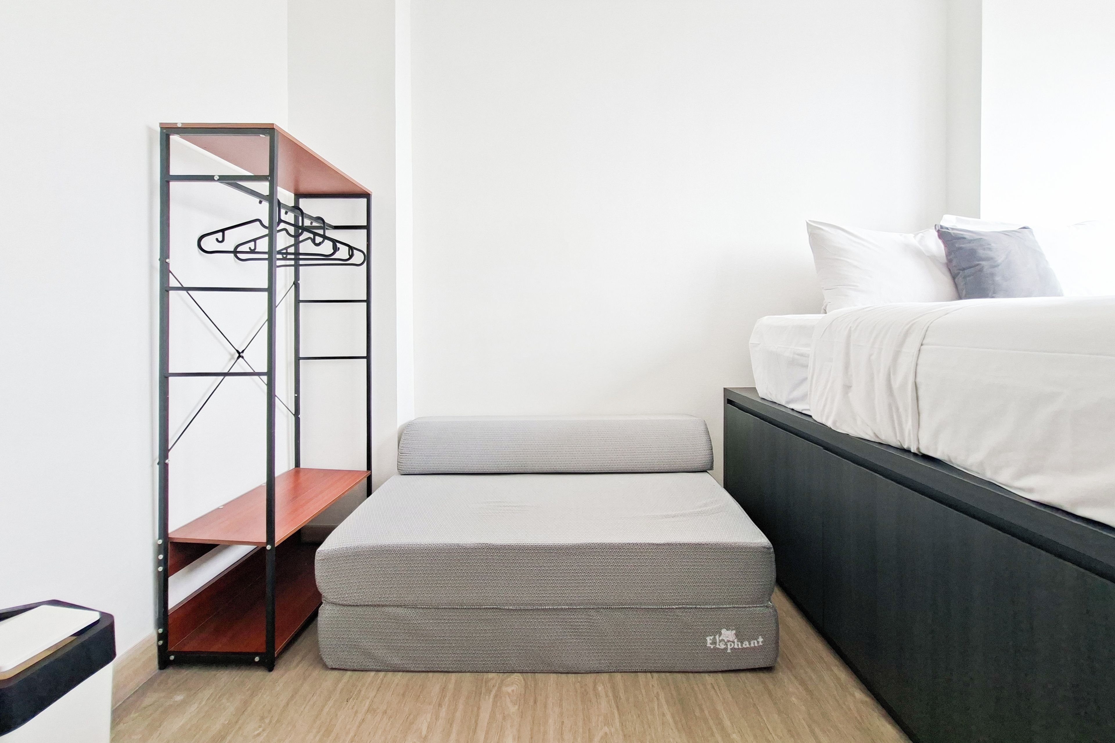 Un dormitorio con cama, un banco de almacenamiento gris y un armario metálico con ropa colgada.