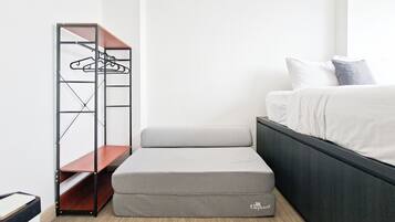 Un dormitorio con cama, un banco de almacenamiento gris y un armario metálico con ropa colgada.