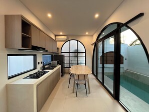 A compact kitchen with a built-in stove, a dining area with a chair, and a large arched window providing a view of the outside. - 2Br Mediterranean Private Pool Villa Nusa Dua (Bali)