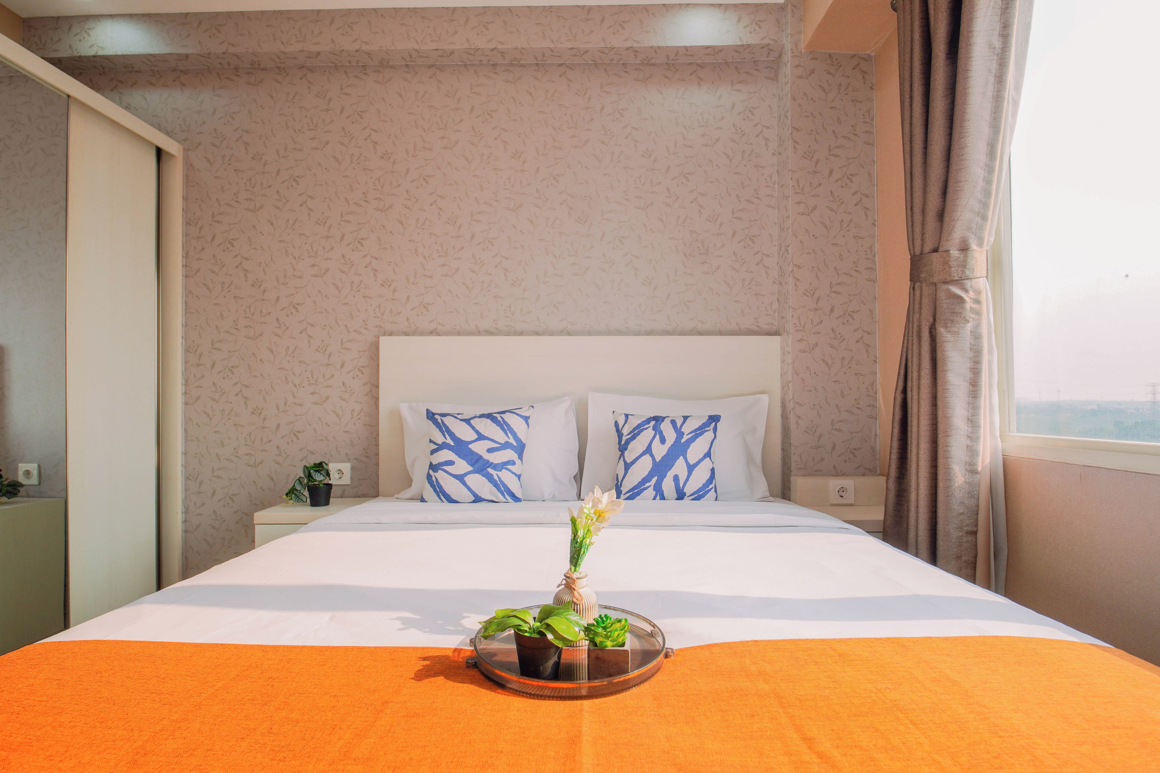 Habitació d'hotel amb llit, dos travessers, una tauleta de nit, una planta en test i una finestra amb cortines.